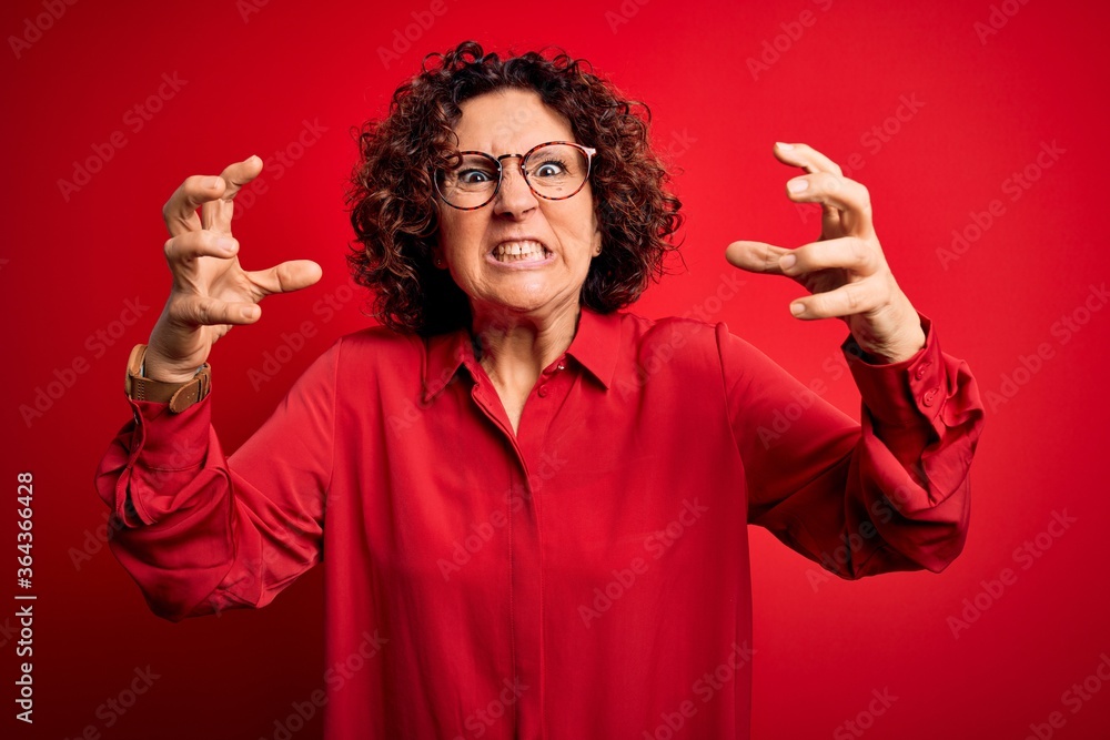 Middle age beautiful curly hair woman wearing casual shirt and glasses over red background Shouting frustrated with rage, hands trying to strangle, yelling mad Middle age beautiful curly hair woman wearing casual shirt and glasses over red background Shouting frustrated with rage, hands trying to strangle, yelling mad