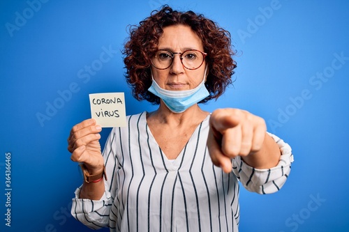 Middle age curly hair woman wearing medical mask holding reminder with corona virus message pointing with finger to the camera and to you  hand sign  positive and confident gesture from the front