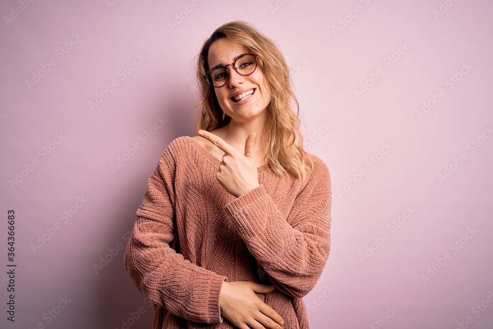 Young beautiful blonde woman wearing casual sweater and glasses over pink background cheerful with a smile of face pointing with hand and finger up to the side with happy and natural expression. Young beautiful blonde woman wearing casual sweater and glasses over pink background cheerful with a smile of face pointing with hand and finger up to the side with happy and natural expression.