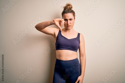 Young beautiful blonde sportswoman doing sport wearing sportswear over white background looking unhappy and angry showing rejection and negative with thumbs down gesture. Bad expression.