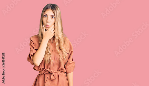 Beautiful caucasian woman with blonde hair wearing summer jumpsuit looking fascinated with disbelief, surprise and amazed expression with hands on chin