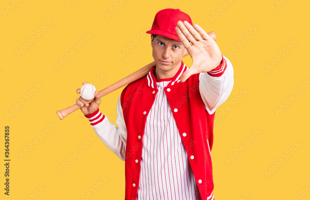 Young handsome man playing baseball holding bat and ball with open hand doing stop sign with serious and confident expression, defense gesture Young handsome man playing baseball holding bat and ball with open hand doing stop sign with serious and confident expression, defense gesture