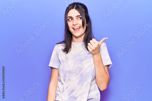 Young beautiful girl wearing casual t shirt pointing thumb up to the side smiling happy with open mouth