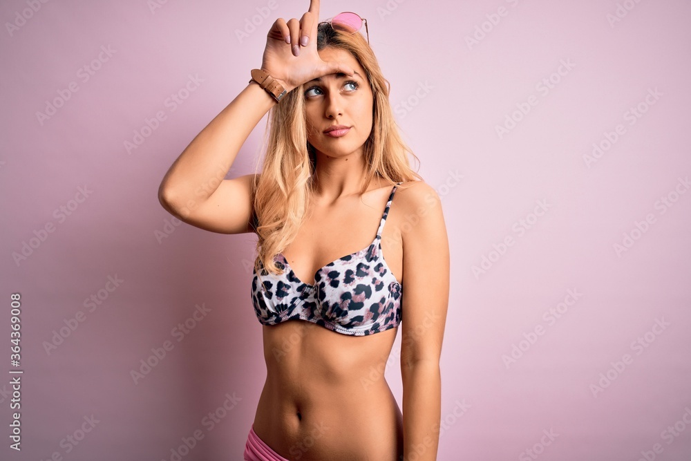 Obraz premium Young beautiful blonde woman on vacation wearing bikini over isolated pink background making fun of people with fingers on forehead doing loser gesture mocking and insulting.