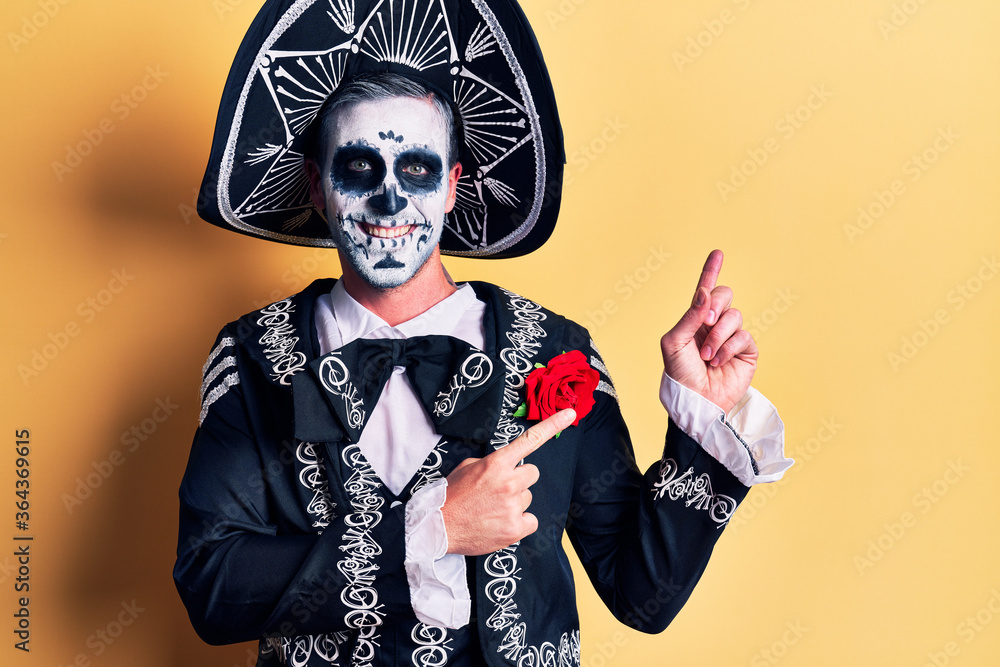 Young man wearing mexican day of the dead costume over yellow smiling and looking at the camera pointing with two hands and fingers to the side.