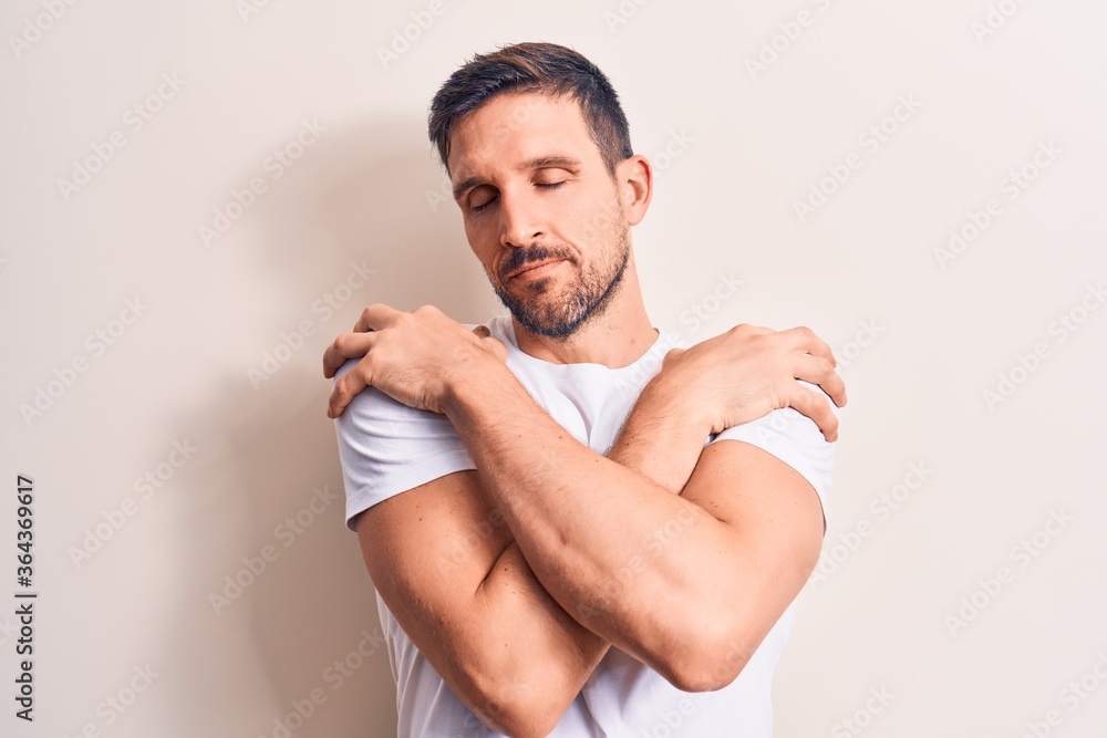 Young handsome man wearing casual t-shirt standing over isolated white background hugging oneself happy and positive, smiling confident. Self love and self care