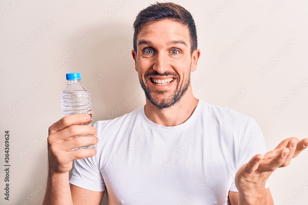 Young handsome man drinking bottle of water to refreshment over isolated white background celebrating achievement with happy smile and winner expression with raised hand Young handsome man drinking bottle of water to refreshment over isolated white background celebrating achievement with happy smile and winner expression with raised hand