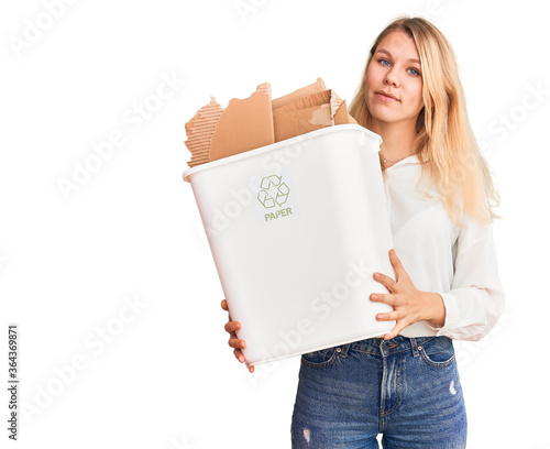 Young beautiful blonde woman holding recycle paper bin thinking attitude and sober expression looking self confident photo