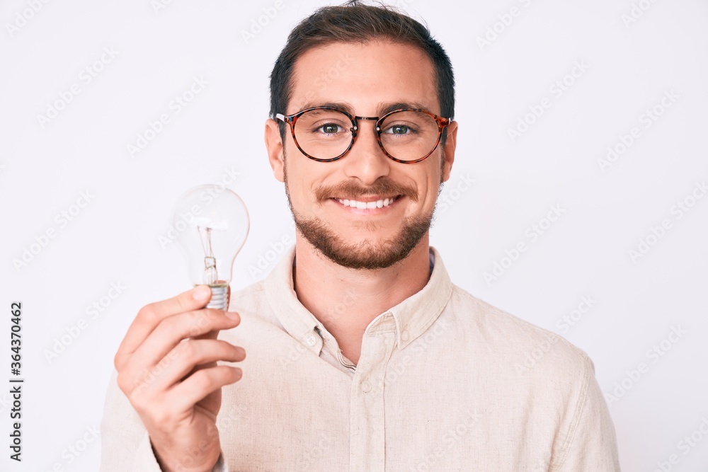 Young handsome man holding lightbulb for inspiration and idea looking positive and happy standing and smiling with a confident smile showing teeth Young handsome man holding lightbulb for inspiration and idea looking positive and happy standing and smiling with a confident smile showing teeth