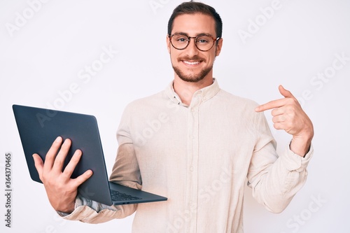 Young handsome man holding laptop pointing finger to one self smiling happy and proud