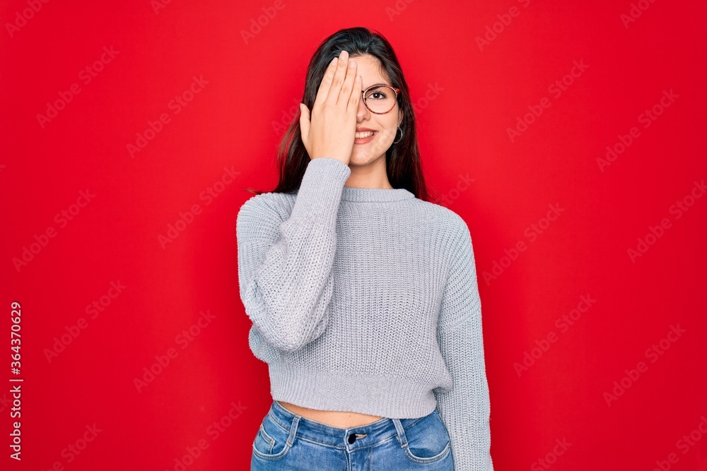 Young beautiful brunette woman wearing casual sweater over red background covering one eye with hand, confident smile on face and surprise emotion. Young beautiful brunette woman wearing casual sweater over red background covering one eye with hand, confident smile on face and surprise emotion.