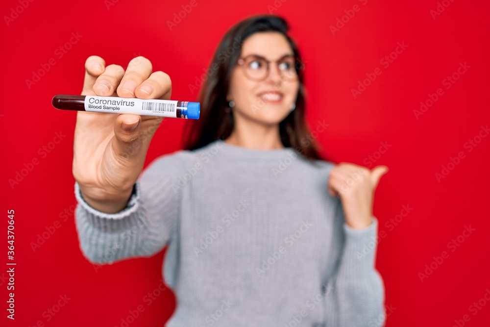 Young beautiful girl wearing glasses holding laboratory blood sample of coronavirus disease pointing and showing with thumb up to the side with happy face smiling Young beautiful girl wearing glasses holding laboratory blood sample of coronavirus disease pointing and showing with thumb up to the side with happy face smiling