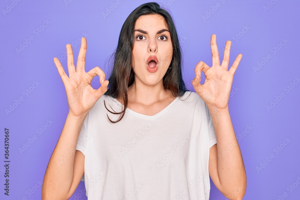 Young beautiful brunette woman wearing casual white t-shirt over purple background looking surprised and shocked doing ok approval symbol with fingers. Crazy expression