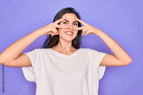 Young beautiful brunette woman wearing casual white t-shirt over purple background Doing peace symbol with fingers over face, smiling cheerful showing victory