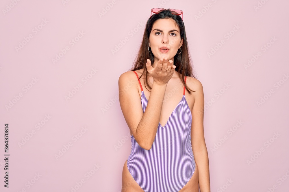 Young beautiful fashion girl wearing swimwear swimsuit and sunglasses over pink background looking at the camera blowing a kiss with hand on air being lovely and sexy. Love expression. Young beautiful fashion girl wearing swimwear swimsuit and sunglasses over pink background looking at the camera blowing a kiss with hand on air being lovely and sexy. Love expression.
