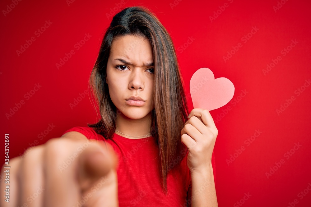 Young beautiful brunette romantic girl holding red paper heart shape over isolated background pointing with finger to the camera and to you, hand sign, positive and confident gesture from the front Young beautiful brunette romantic girl holding red paper heart shape over isolated background pointing with finger to the camera and to you, hand sign, positive and confident gesture from the front