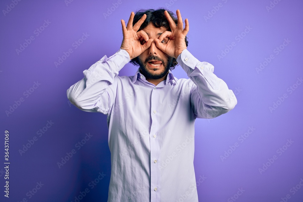 Young handsome business man with beard wearing shirt standing over purple background doing ok gesture like binoculars sticking tongue out, eyes looking through fingers. Crazy expression.