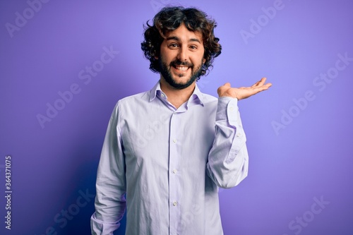 Young handsome business man with beard wearing shirt standing over purple background smiling cheerful presenting and pointing with palm of hand looking at the camera.