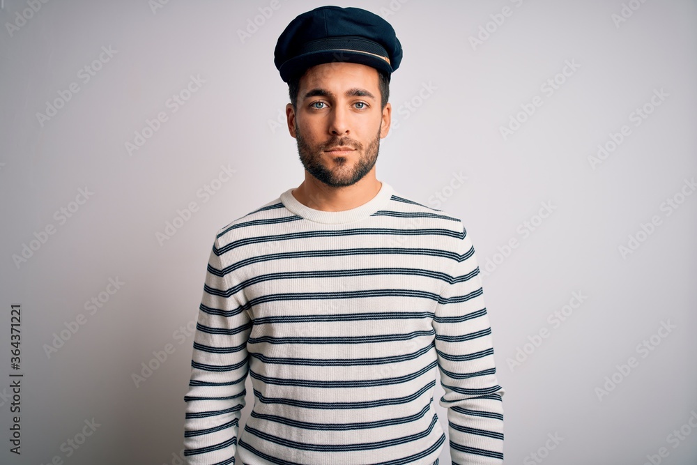 Young handsome sailor man with beard wearing navy striped uniform and captain hat with serious expression on face. Simple and natural looking at the camera.
