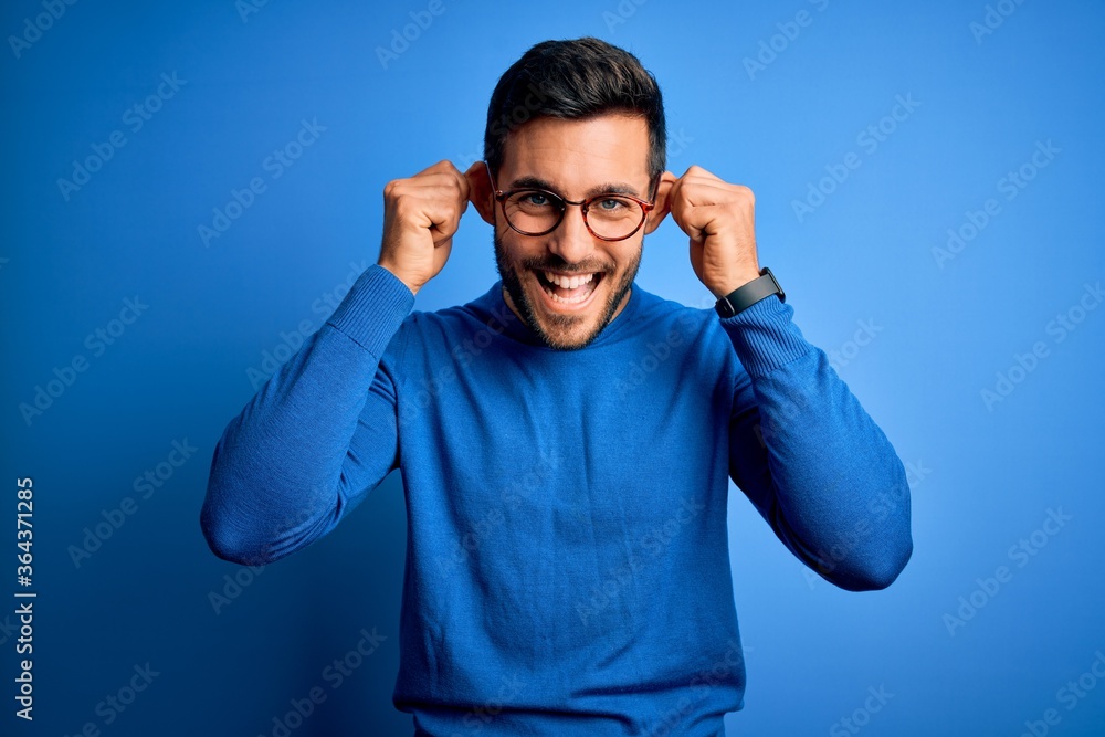 Young handsome man with beard wearing casual sweater and glasses over blue background Smiling pulling ears with fingers, funny gesture. Audition problem Young handsome man with beard wearing casual sweater and glasses over blue background Smiling pulling ears with fingers, funny gesture. Audition problem