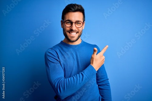 Young handsome man with beard wearing casual sweater and glasses over blue background cheerful with a smile on face pointing with hand and finger up to the side with happy and natural expression photo