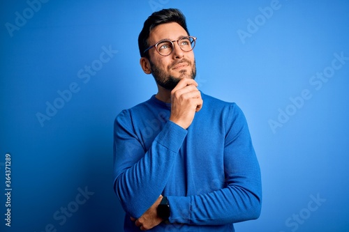 Young handsome man with beard wearing casual sweater and glasses over blue background with hand on chin thinking about question, pensive expression. Smiling and thoughtful face. Doubt concept.