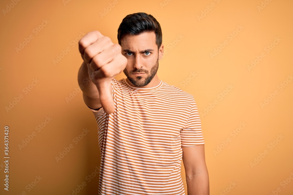 Young handsome man with beard wearing casual striped t-shirt over yellow background looking unhappy and angry showing rejection and negative with thumbs down gesture. Bad expression. Young handsome man with beard wearing casual striped t-shirt over yellow background looking unhappy and angry showing rejection and negative with thumbs down gesture. Bad expression.