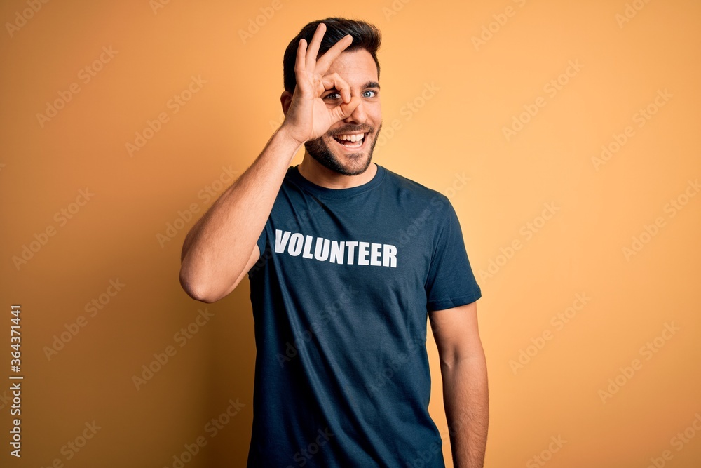 Young handsome man with beard volunteering wearing t-shirt with volunteer message with happy face smiling doing ok sign with hand on eye looking through fingers Young handsome man with beard volunteering wearing t-shirt with volunteer message with happy face smiling doing ok sign with hand on eye looking through fingers