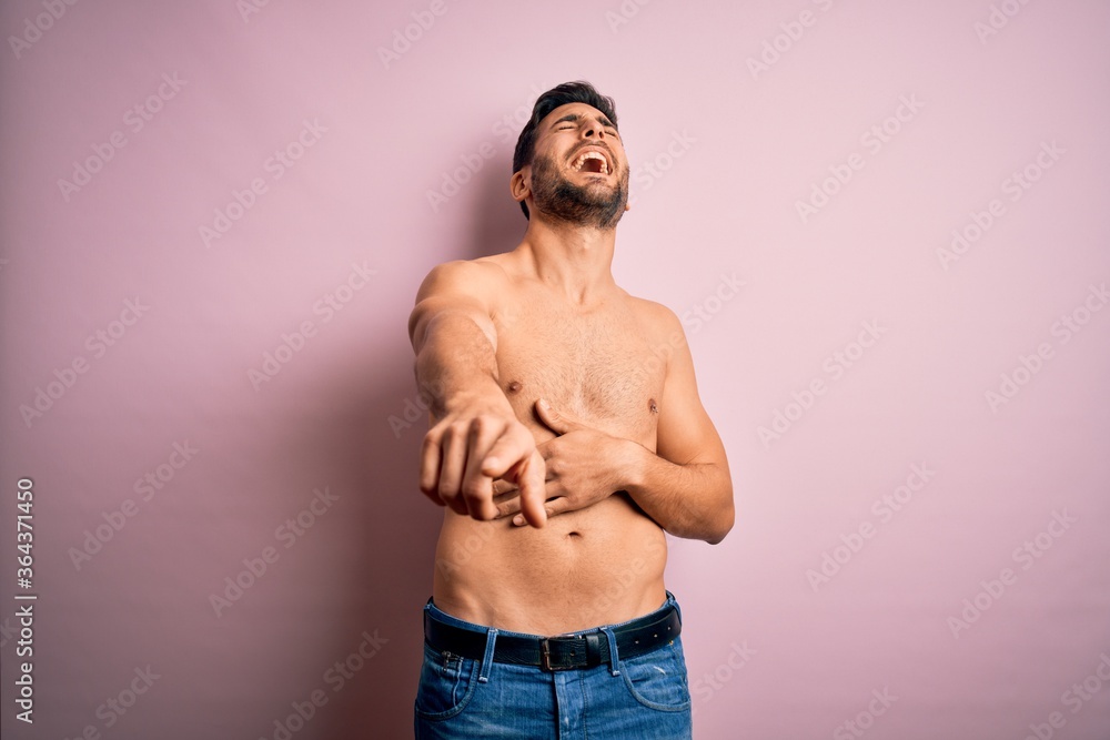 Young handsome strong man with beard shirtless standing over isolated pink background laughing at you, pointing finger to the camera with hand over body, shame expression Young handsome strong man with beard shirtless standing over isolated pink background laughing at you, pointing finger to the camera with hand over body, shame expression