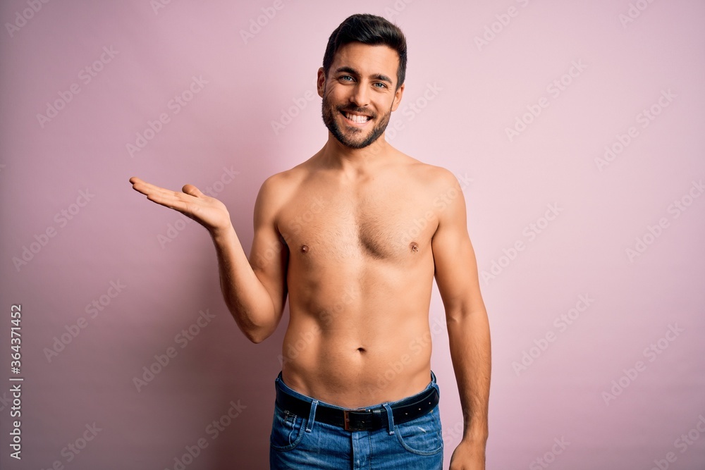 Young handsome strong man with beard shirtless standing over isolated pink background smiling cheerful presenting and pointing with palm of hand looking at the camera.