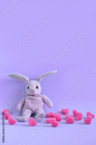 Pink plush hare sits on violet background among soft pink hearts, vertical orientation. Valentine's day card