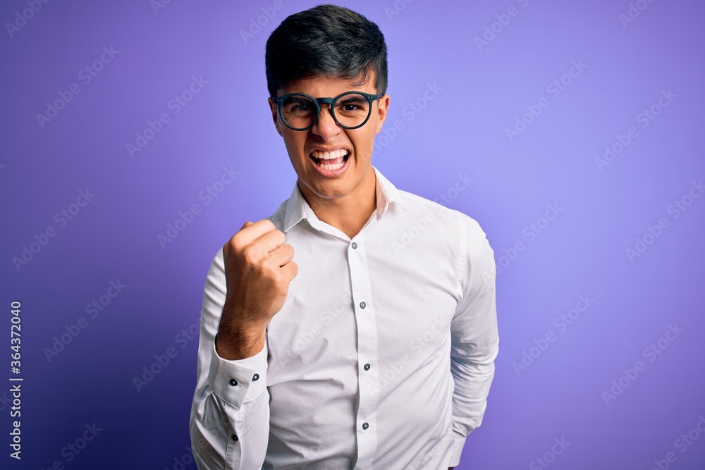 Young handsome business man wearing shirt and glasses over isolated purple background angry and mad raising fist frustrated and furious while shouting with anger. Rage and aggressive concept. Young handsome business man wearing shirt and glasses over isolated purple background angry and mad raising fist frustrated and furious while shouting with anger. Rage and aggressive concept.