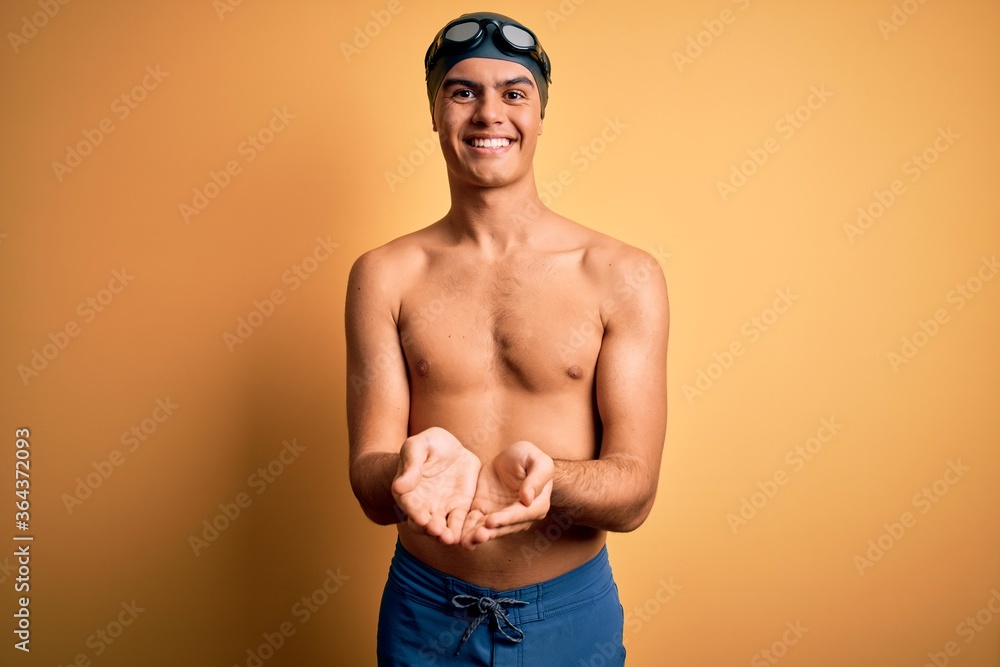 Young handsome man shirtless wearing swimsuit and swim cap over isolated yellow background Smiling with hands palms together receiving or giving gesture. Hold and protection