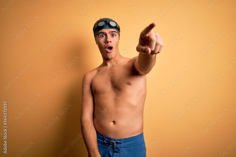 Young handsome man shirtless wearing swimsuit and swim cap over isolated yellow background Pointing with finger surprised ahead, open mouth amazed expression, something on the front