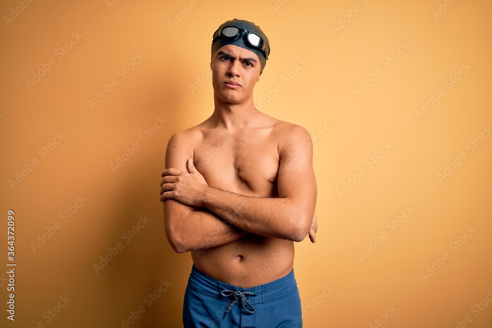 Young handsome man shirtless wearing swimsuit and swim cap over isolated yellow background skeptic and nervous, disapproving expression on face with crossed arms. Negative person.