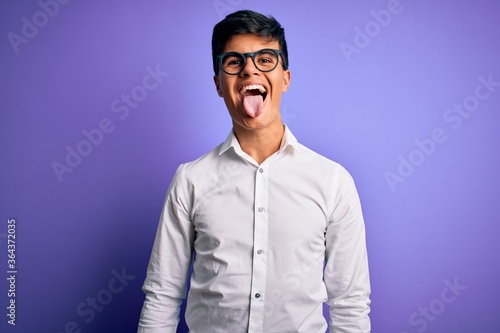 Young handsome business man wearing shirt and glasses over isolated purple background sticking tongue out happy with funny expression. Emotion concept.