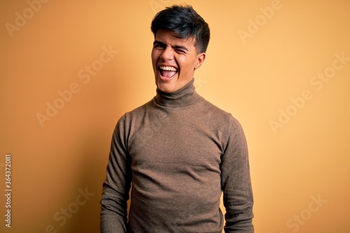 Young handsome man wearing casual turtleneck sweater over isolated yellow background winking looking at the camera with sexy expression, cheerful and happy face. © Krakenimages.com