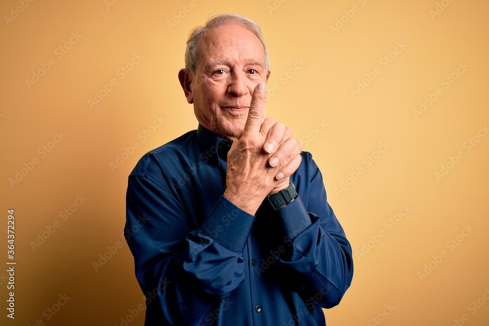 Grey haired senior man wearing casual blue shirt standing over yellow background Holding symbolic gun with hand gesture, playing killing shooting weapons, angry face Grey haired senior man wearing casual blue shirt standing over yellow background Holding symbolic gun with hand gesture, playing killing shooting weapons, angry face
