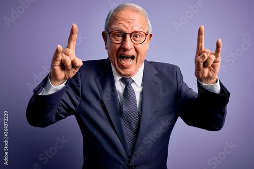 Grey haired senior business man wearing glasses and elegant suit and tie over purple background shouting with crazy expression doing rock symbol with hands up. Music star. Heavy music concept.