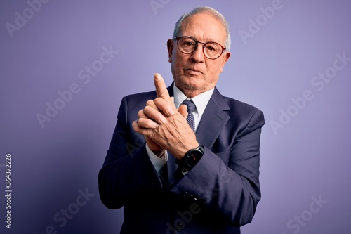 Grey haired senior business man wearing glasses and elegant suit and tie over purple background Holding symbolic gun with hand gesture, playing killing shooting weapons, angry face