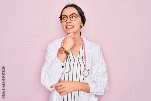 Beautiful doctor woman with blue eyes wearing coat and stethoscope over pink background with hand on chin thinking about question  pensive expression. Smiling with thoughtful face. Doubt concept.
