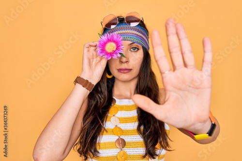Young beautiful hippie woman with blue eyes wearing sunnglasses holding pink flower over eye with open hand doing stop sign with serious and confident expression, defense gesture © Krakenimages.com