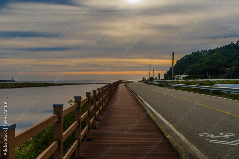 Wooden walkway along a country road next to waterway with beautiful sunset over the ocean in the background.