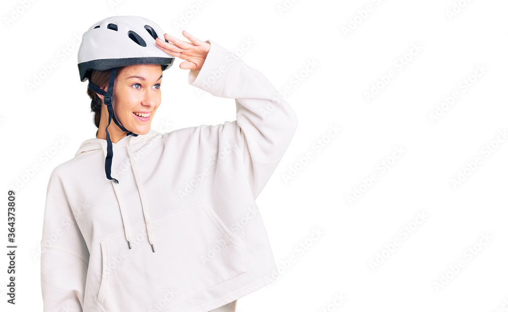 Beautiful brunette young woman wearing bike helmet and sporty clothes very happy and smiling looking far away with hand over head. searching concept. Beautiful brunette young woman wearing bike helmet and sporty clothes very happy and smiling looking far away with hand over head. searching concept.