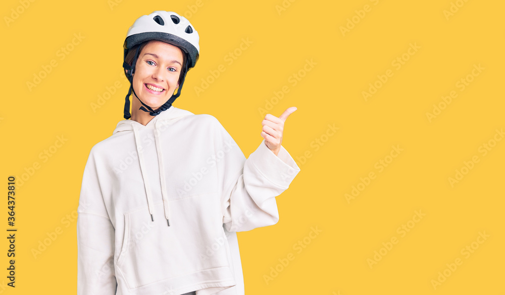 Beautiful brunette young woman wearing bike helmet and sporty clothes smiling with happy face looking and pointing to the side with thumb up.