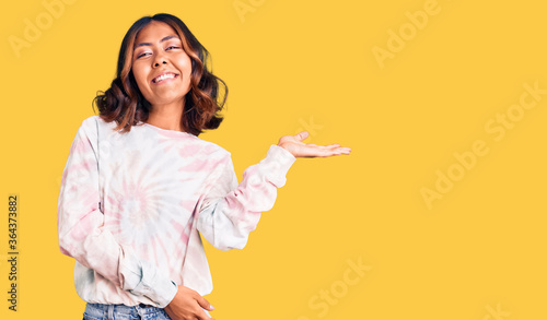 Young beautiful mixed race woman wearing casual tie dye sweatshirt smiling cheerful presenting and pointing with palm of hand looking at the camera.