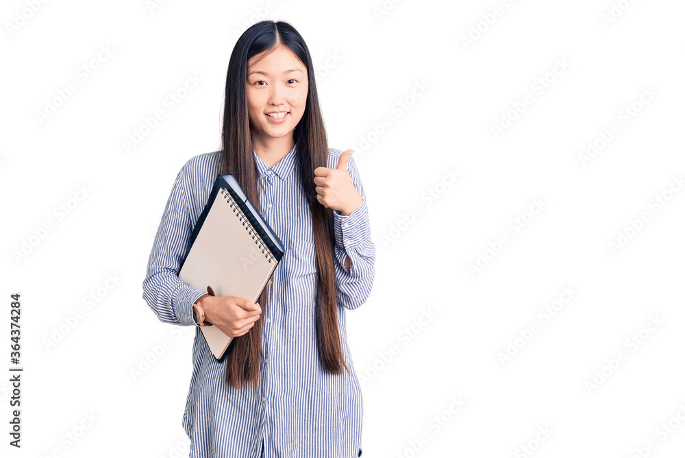 Young beautiful chinese woman holding notebook smiling happy and positive, thumb up doing excellent and approval sign Young beautiful chinese woman holding notebook smiling happy and positive, thumb up doing excellent and approval sign