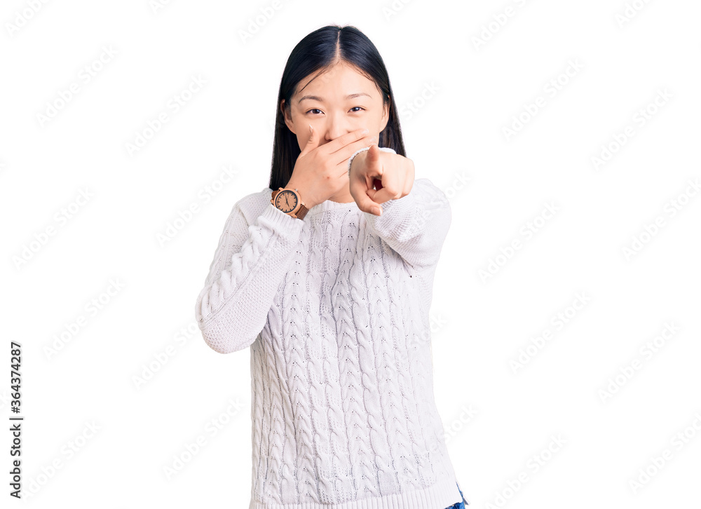 Young beautiful chinese woman wearing casual sweater laughing at you, pointing finger to the camera with hand over mouth, shame expression
