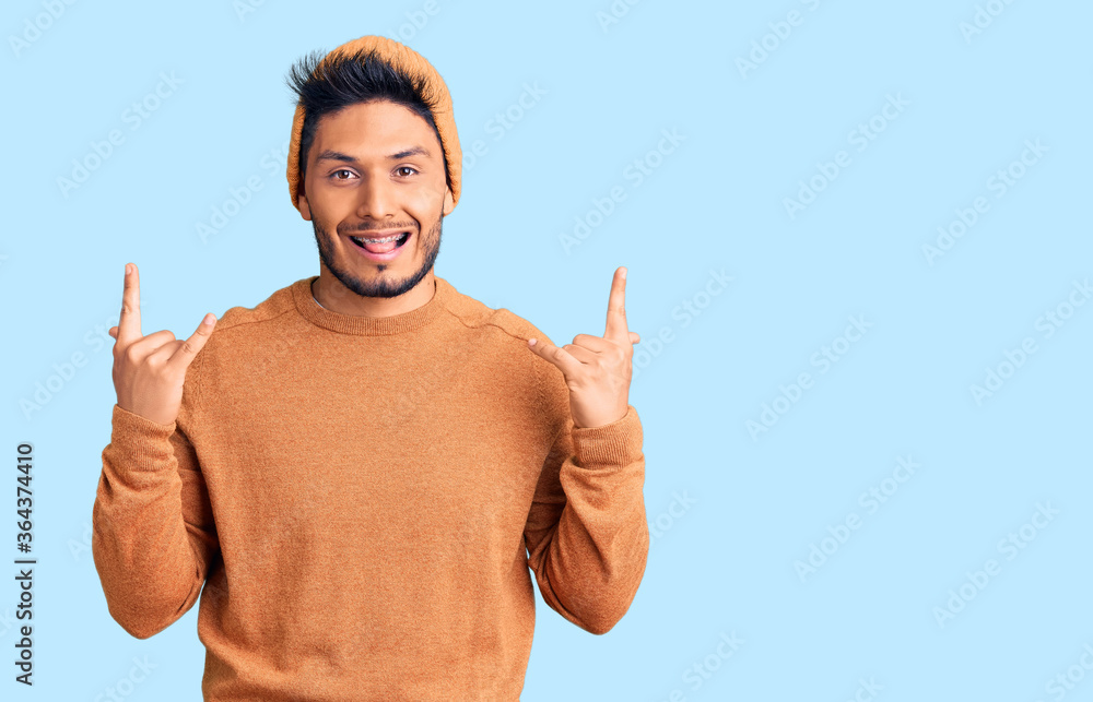 Handsome latin american young man wearing winter sweater and wool hat shouting with crazy expression doing rock symbol with hands up. music star. heavy concept. Handsome latin american young man wearing winter sweater and wool hat shouting with crazy expression doing rock symbol with hands up. music star. heavy concept.