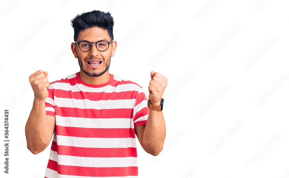 Handsome latin american young man wearing casual clothes and glasses angry and mad raising fists frustrated and furious while shouting with anger. rage and aggressive concept.